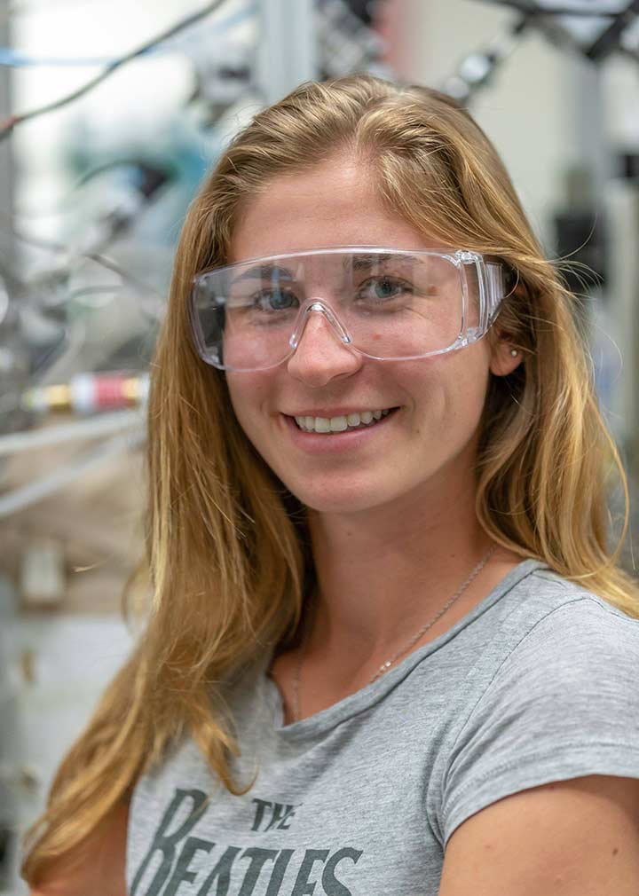 A woman wearing safety goggles smiles in front of a research laboratory