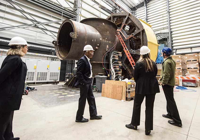 Four people in hard hats, inside a building, stand next to a large dynometer device.