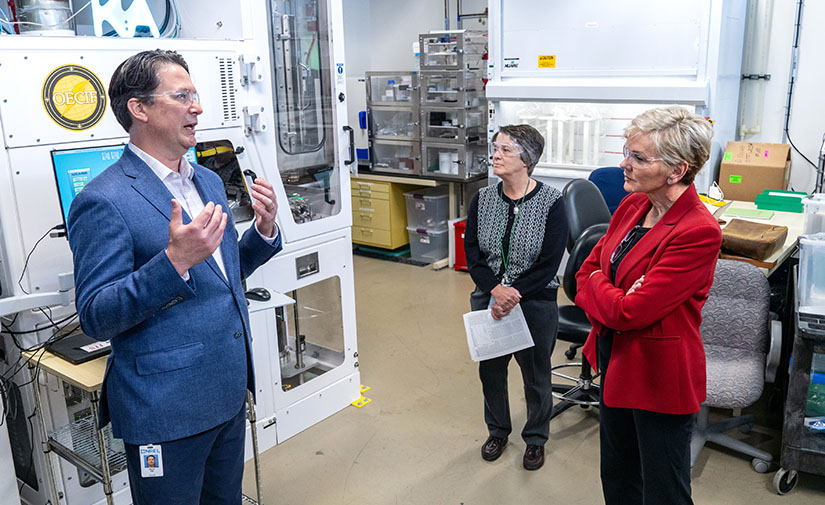 A man standing in front of a piece of a scientific equipment talks to two women.