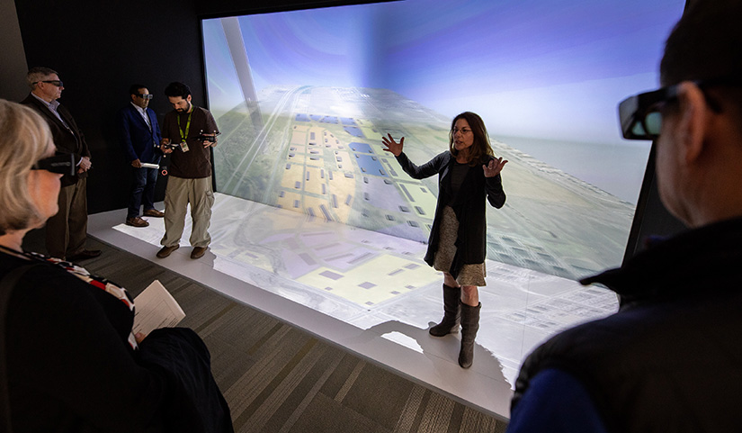 A person stands in front of a screen speaking to a group wearing 3D glasses.
