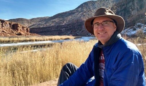 A man sits on a river bank with cliffs in the background.