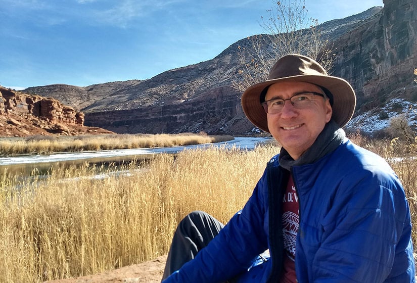 20250505-nrel-researcher-craig-turchi-brings-small-business-experience-to-big-concentrating-solar-projects-river A man sits on a river bank with cliffs in the background.