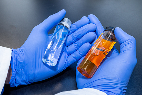 20250520-nrel-crysalis-biosciences-collaborate-to-scale-up-domestic-biomanufacturing-technologies-98211 Two gloved hands hold a pair of vials, one with clear liquid and one with amber liquid.