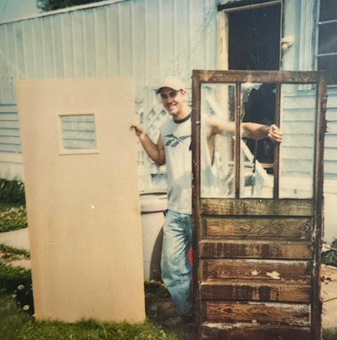 20250701-built-on-helping-others-cory-chovanec-journey-weatherization-dresser A person standing in between a new door and an old door.