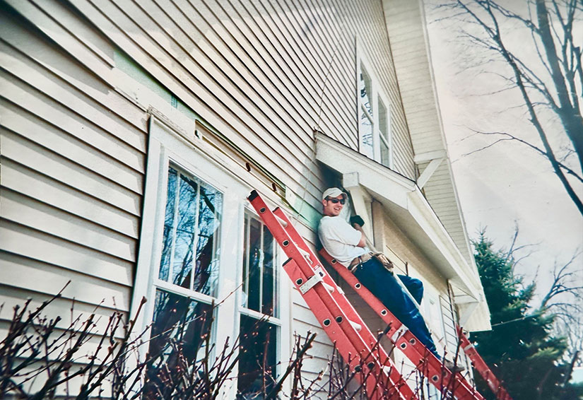 20250701-built-on-helping-others-cory-chovanec-journey-weatherization-ladder A person stands on a ladder leaning against a house.