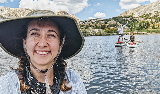A person wearing a hat taking a selfie in front of two paddleboarders on a body of water.