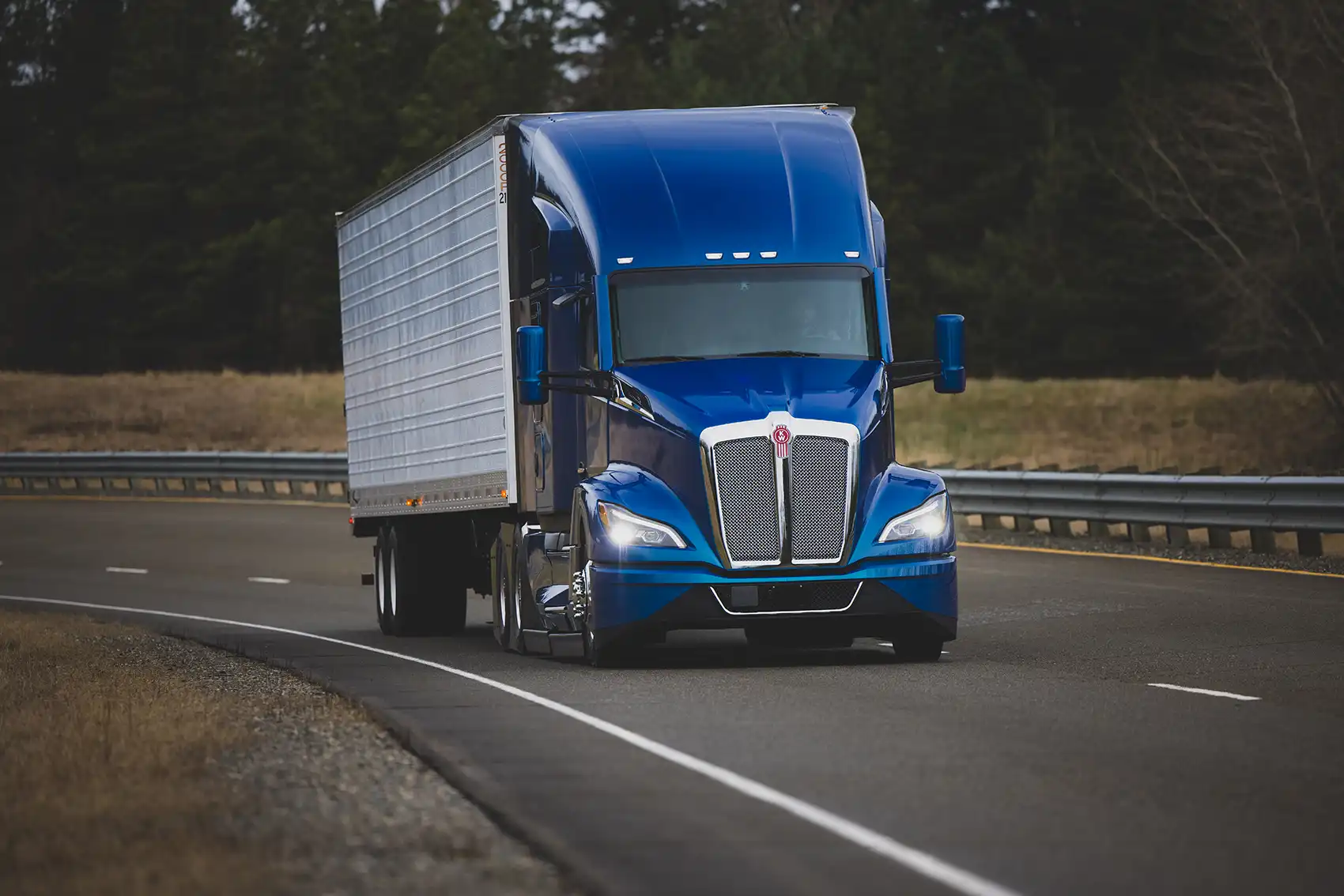 20250915-nrel-expertise-contributes-customizable-ready-deploy-tool-package-improve-fleet-efficiency-freight A blue freight truck in the foreground on a highway flanked by grass and dark trees in the background.