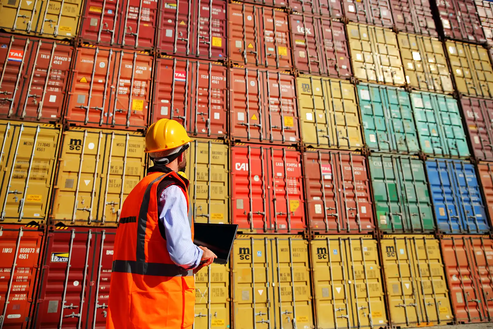 20251118-nrel-informes-tool-will-enable-streamlined-cost-effective-intermodal-freight-getty A man in a hard hat and safety vest inventories a tall, colorful stack of standardized metal shipping containers.