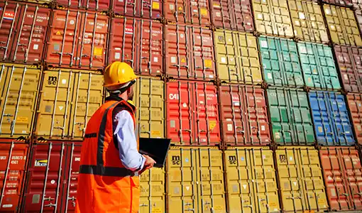 Continue reading about A man in a hard hat and safety vest inventories a tall, colorful stack of standardized metal shipping containers.