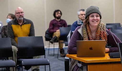 A woman sitting at a desk with a laptop and microphone to present while an audience behind her watches.