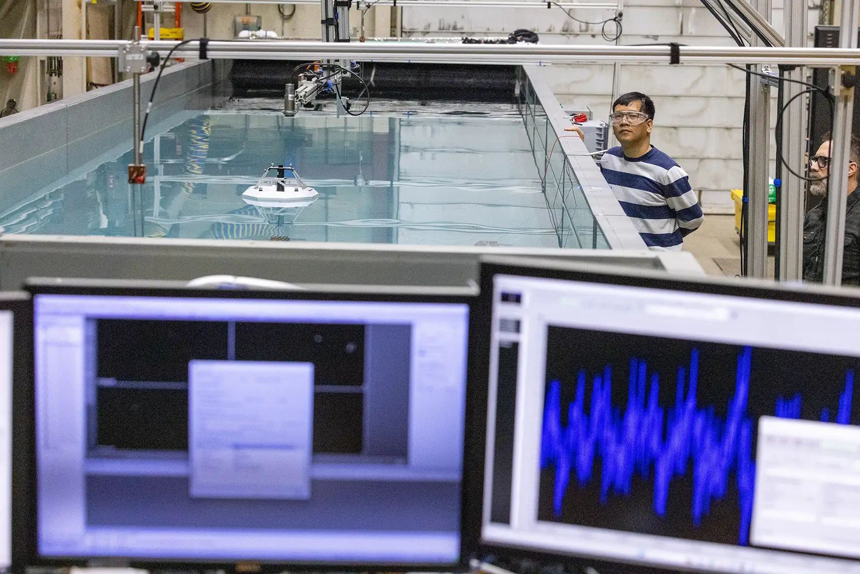 20260112-from-beta-to-data-marine-energy-analysis-tool-is-now-stable-and-ready-98259 A person wearing safety goggles stands next to a large tank of water with a frisbee-sized device floating inside and computer screens visible in the foreground.
