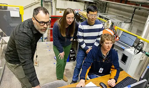 Four people stand or sit while looking at three computer monitors.