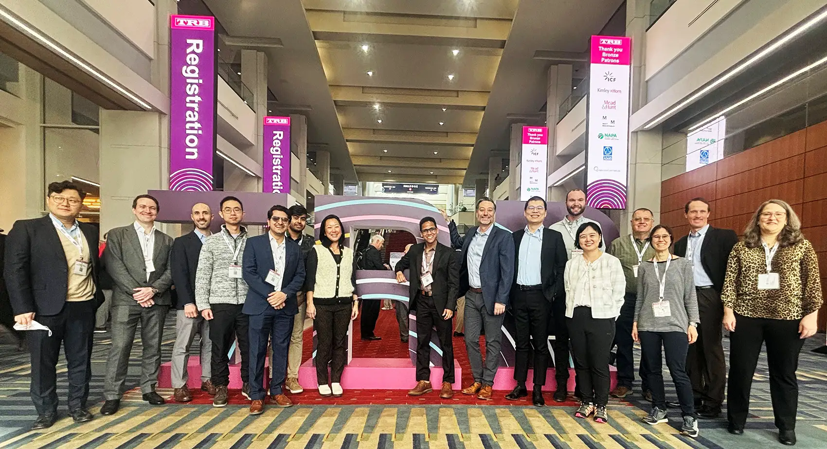 Sixteen people posing in front of TRB sign in large lobby.