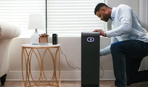 A man in a blue shirt and jeans plugs a black smart battery into his home Wi-Fi router, beside a white couch, with a plant, lamp, and window with blinds in the background.