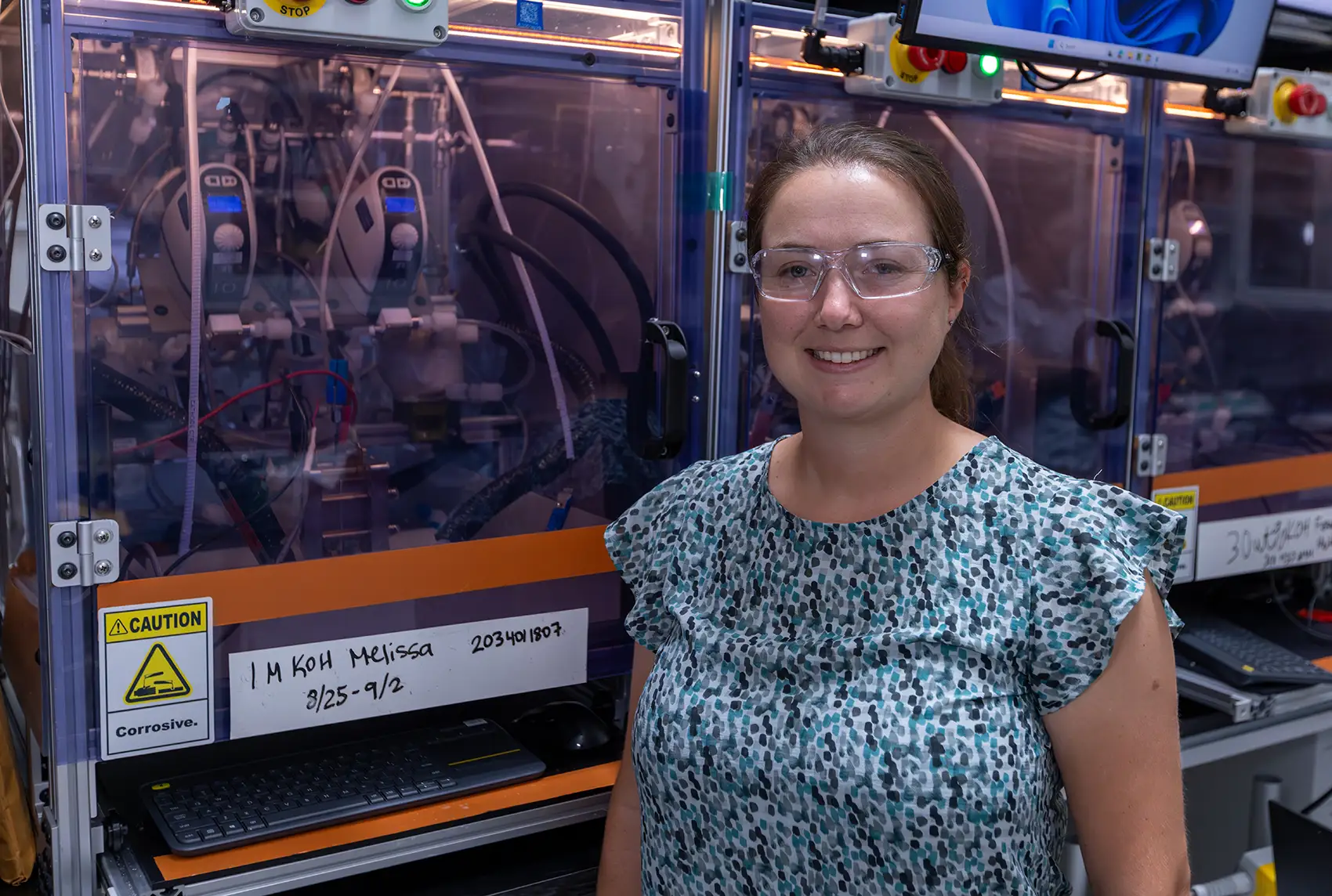 20260127-early-career-spotlight-from-coastal-curiosity-to-catalyst-discovery-how-melissa-kreider-found-her-element-101806 A woman with safety goggles stands in front of electrolyzer testing equipment.