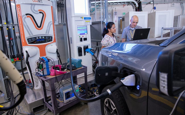 transportation-mobility-82750 Two people wearing eye protection look at a laptop propped on the hood of an electric vehicle as it charges inside a laboratory.
