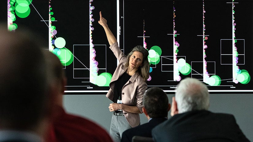 Person pointing up to data displayed on large screen in front of conference room