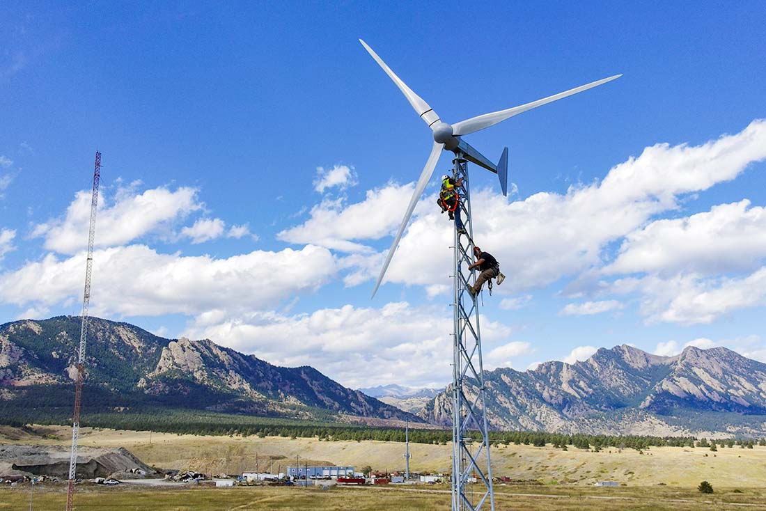 dswind-bergey-windpower-excel-1-5 Two workers wearing protective gear work near the top of a small wind turbine with mountains in the background.