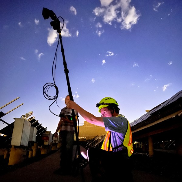 A man in a hard hat and safety vest holds a tall piece of equipment up behind some solar panels. Another man takes notes in the background.