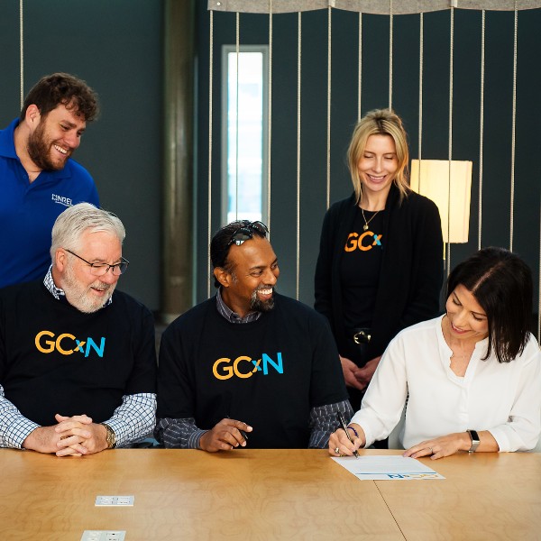 Five people--three seated at a table and two standing--watch another person as they sign. Three of the people wear shirts that read GCxN.