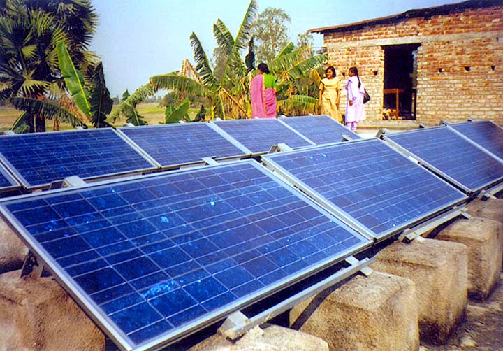 Three women standing next to a ground-mounted solar photovoltaic system mounted on blocks in front of a brick building.