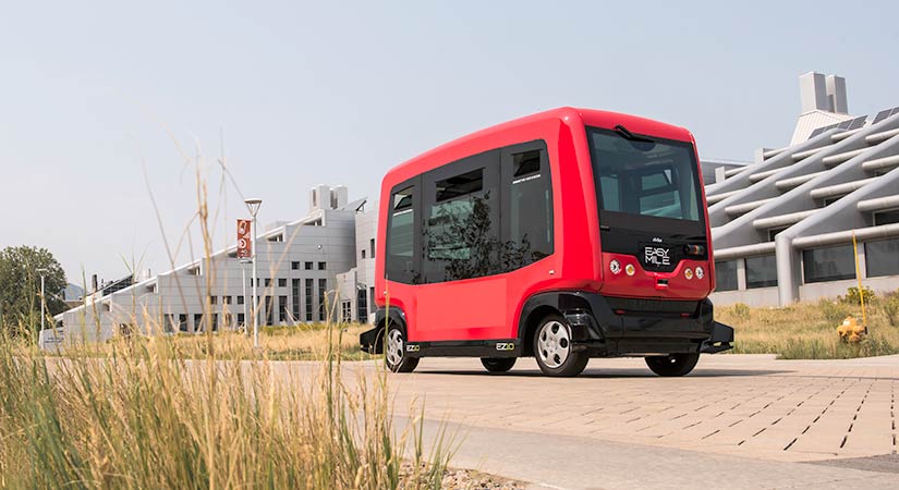 A small, red, square-like all-electric vehicle in front of a white building with grass in the foreground.