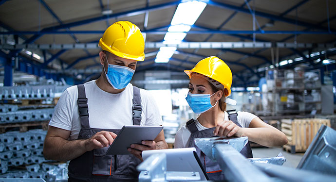 Workers in hardhats in a warehouse with steel parts; both workers are looking at a handheld device that one of the workers is holding.