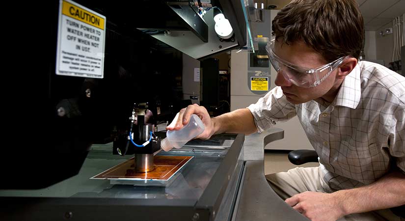 A photo of a researcher wearing protective eyewear as he squirts a clear liquid onto a tray under machinery.