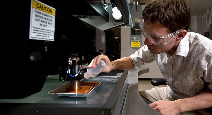 A photo of a researcher wearing protective eyewear as he squirts a clear liquid onto a tray under machinery.