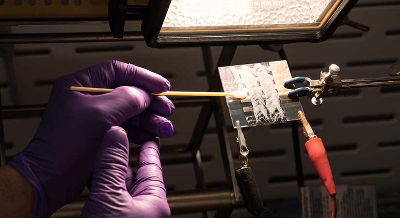 Gloved hands use cotton swab on a square perovskite solar cell under a light.