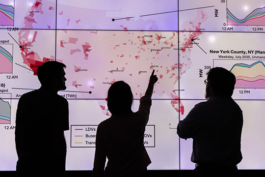 202408-evi-x-89546 Three silhouetted people looking at a data visualization with lines, charts, and a U.S. map on screens behind them