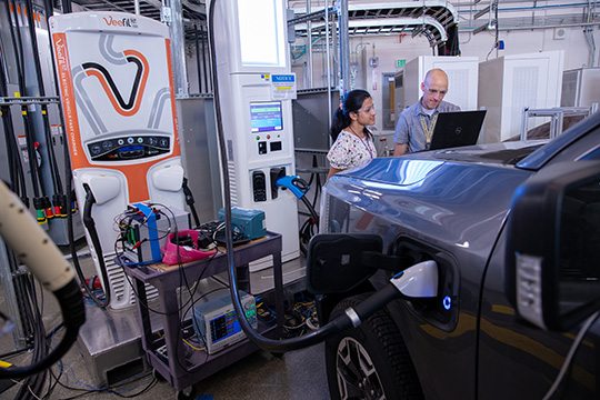 202408-nextgen-profiles Two people look at a laptop placed on the hood of an EV plugged into a charger in an indoor NREL lab.