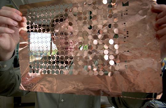 202408-short-circuiting-on-purpose-how-nrel-licensed-tech-continues-to-make-difference-in-space-34831 A person holds a sheet of copper discs in front of their face. Some of the sheet has empty holes where the discs have already come out.