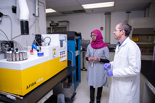 202408-sustainable-vehicle-fuels-70453 Two people in a fuels and combustion lab at NREL looking at a rectangular fuel analysis machine.