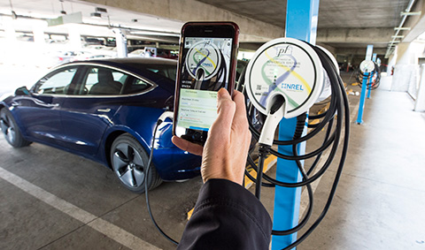 A hand holding a phone with a charging application running on it, in front of an electric vehicle and a charging station in a parking garage.