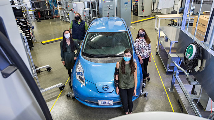 Four people wearing masks stand in a circle around an electric vehicle.