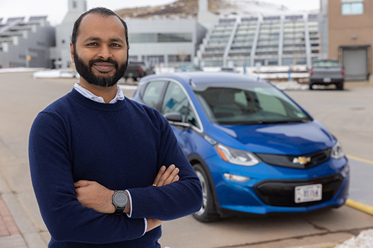 fall24-88010 Ranjit Desai, smiling with arms folded in front of a parked light-duty passenger EV.