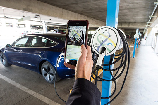fall24-scm A person's hand holding a smartphone showing a smart charge management app in front of a plugged-in blue electric car.