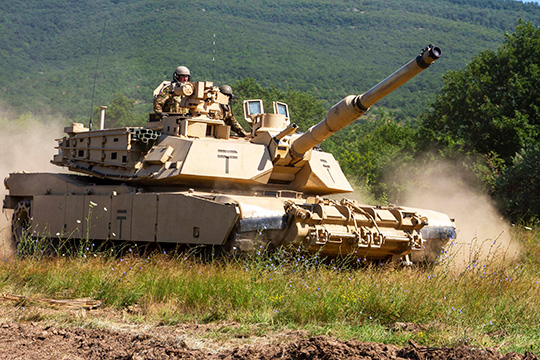 fall24-tank Several U.S. soldiers driving an M1 Abrams tank across a green field, kicking up a cloud of dust.