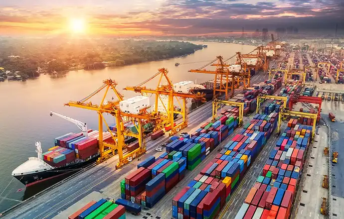 A busy port scene at sunrise. Cranes unload containers from cargo ships and stack them in colorful rows.