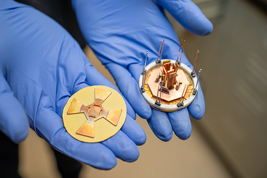 Two gloved hands hold a small octagonal power module and a corresponding attachment layer.