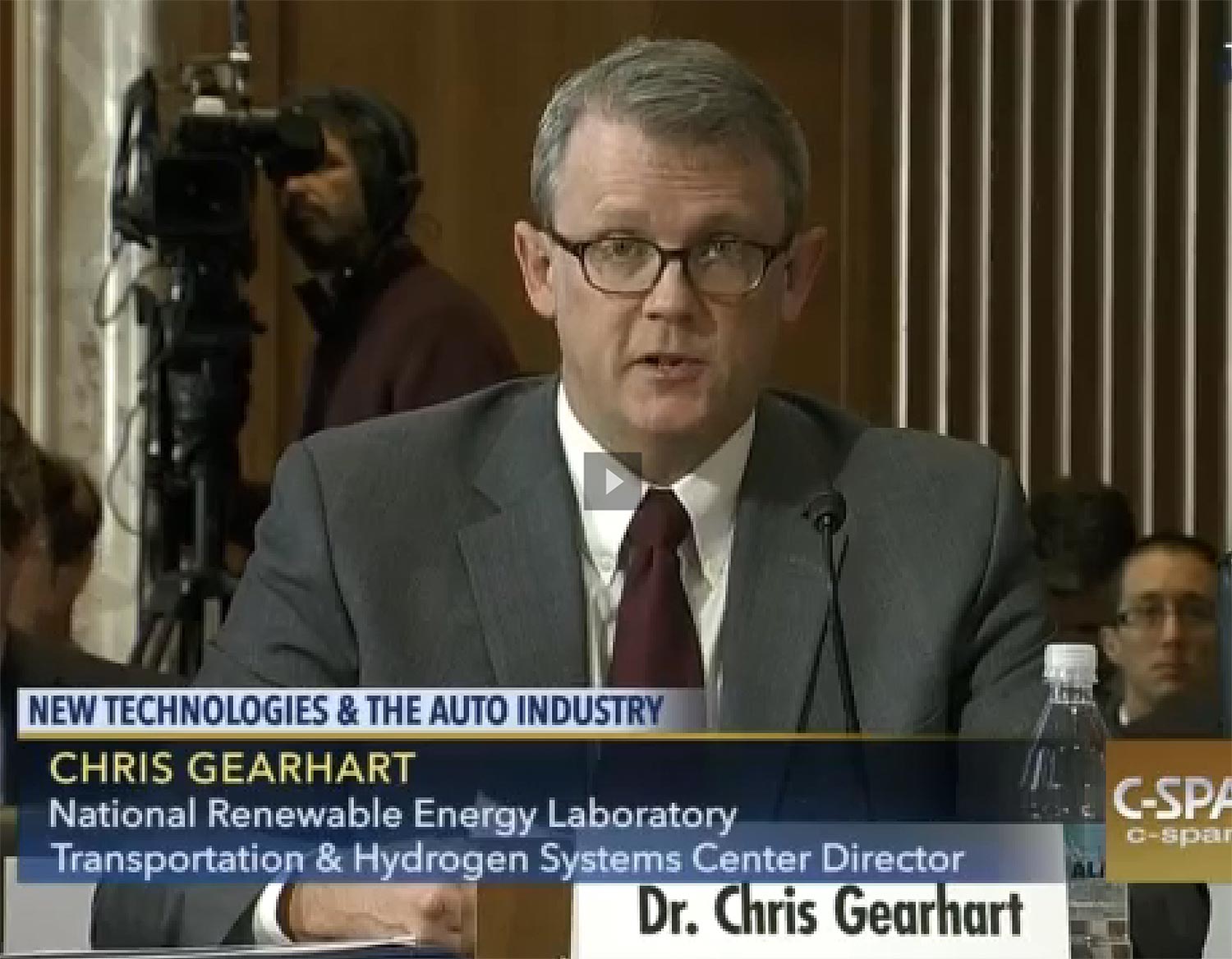 Photo of a man wearing glasses and in a suit sitting in front of a nameplate that says: "Dr. Chris Gearhart." Text over image says: "New Technologies & The Auto Industry; Chris Gearhart, National Renewable Energy Laboratory, Transportation Systems Center Director.