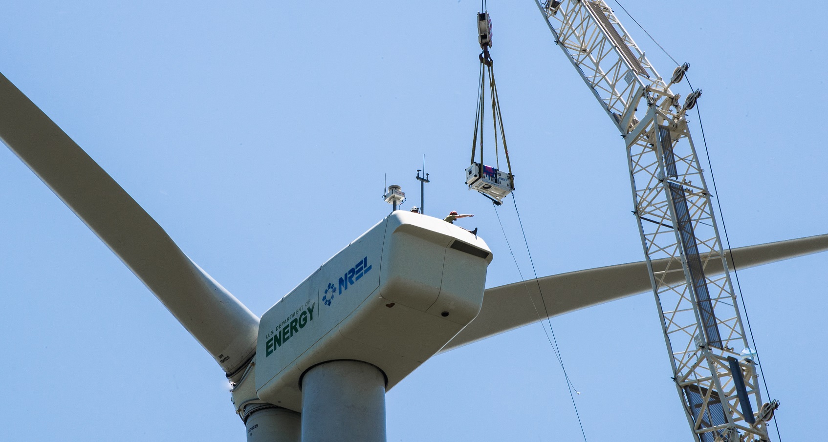 Photo of the top of a turbine and a crane lowering a piece of hardware onto the nacelle.