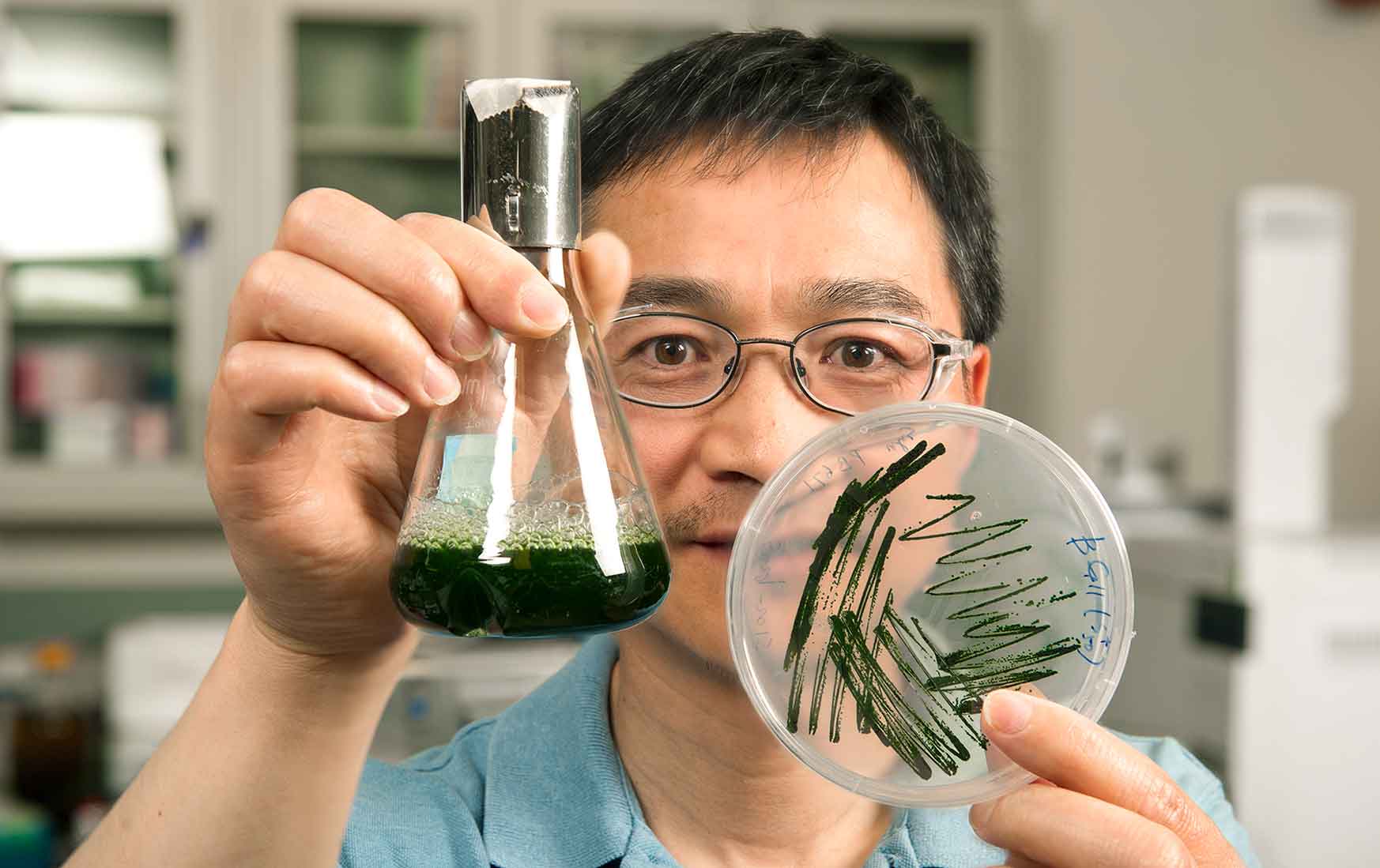 Photo of a man holding a beaker of green liquid and a petri dish with green growth.