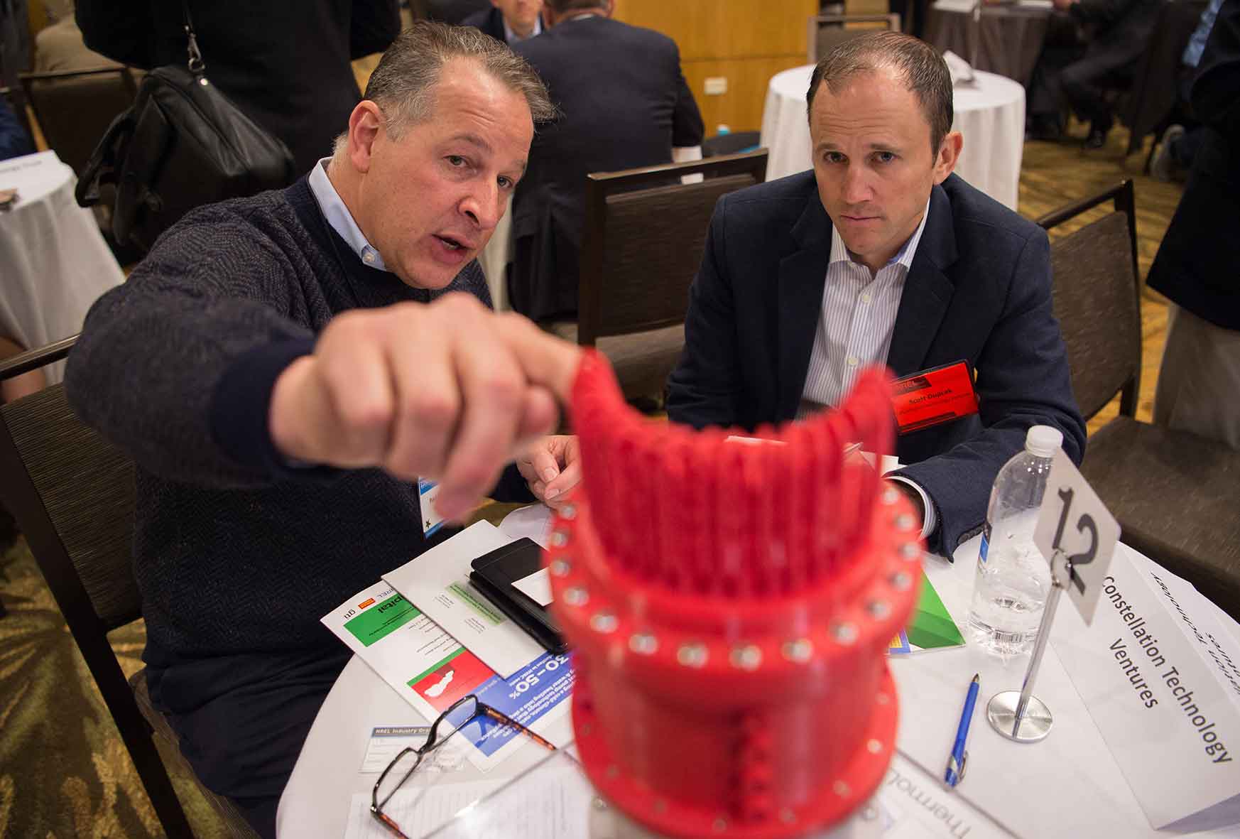 A man points to a device while talking to a man seated next to him.