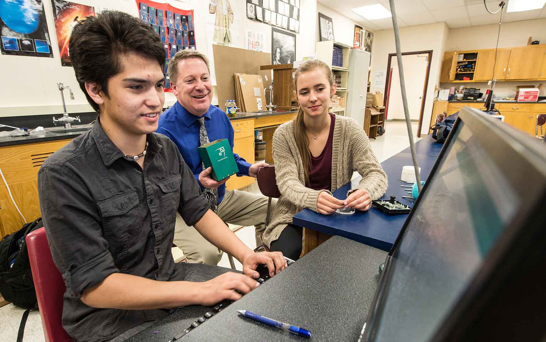 Photo shows a male student working on a computer while a teacher and another student watch.