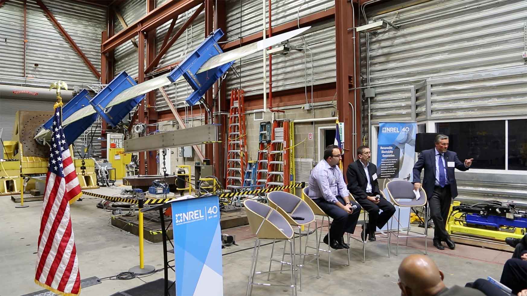 A man stands and talks as two others sit on a podium in an equipment warehouse.