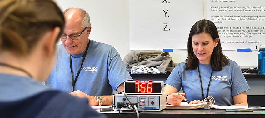 Two NREL staff members, one female on the right and one male on the left, sit at a desk reading questions and judging answers during a match at the 2018 High School Science Bowl.