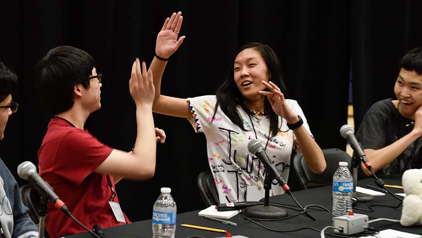 Two teammates, one girl and one boy, high-five after a correct answer while two other boys on their team look on and smile.  