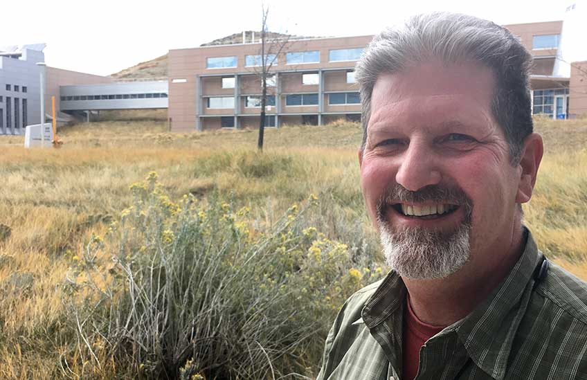 Photo of a man standing at the base of a hill covered by long brown and green grasses, topped by a large modern facility.